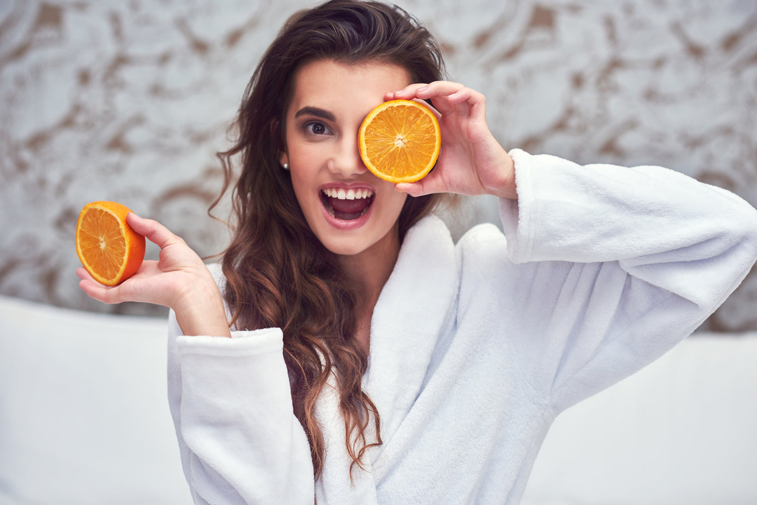 Woman wearing a robe holding an orange cut into two pieces, holding one half over her eye.